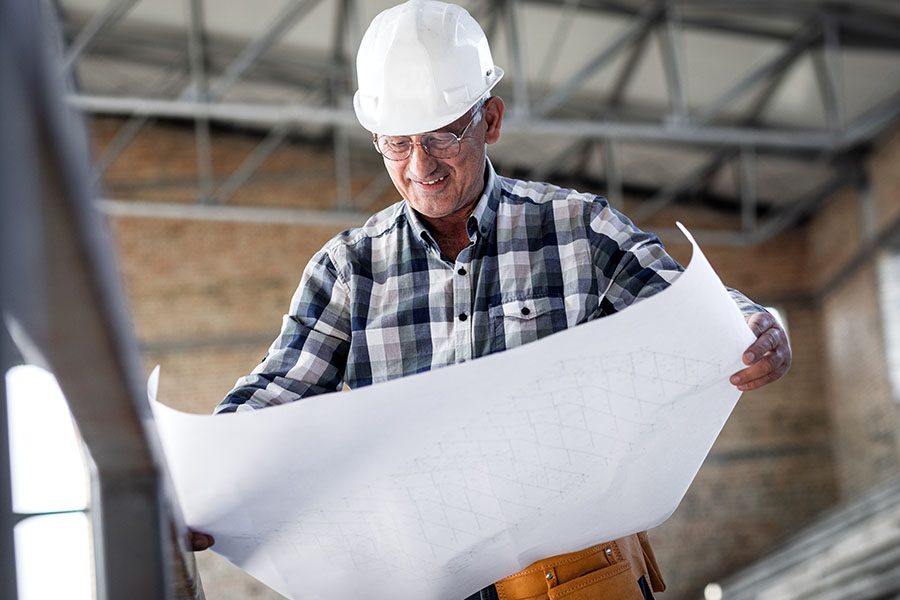 Specialized Business Insurance - Portrait of a Smiling Mature Contractor Standing Inside a New Building Construction Worksite While Looking at Building Blueprints